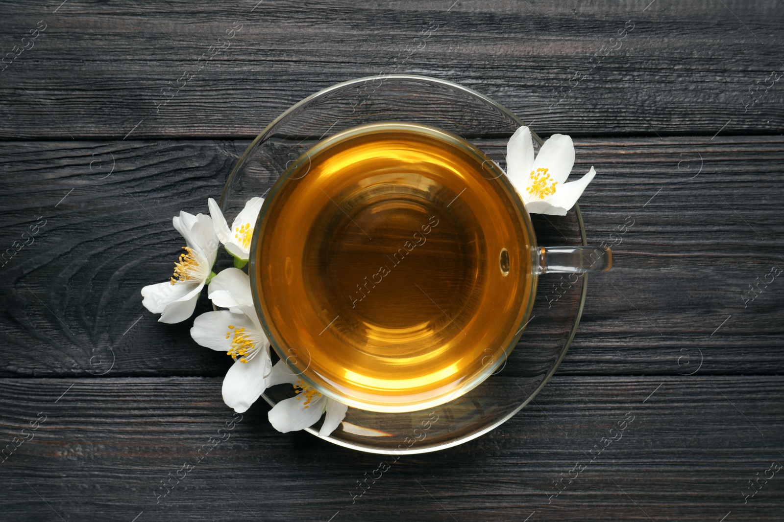 Photo of Glass cup of aromatic jasmine tea and fresh flowers on black wooden table, top view