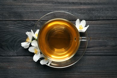 Glass cup of aromatic jasmine tea and fresh flowers on black wooden table, top view