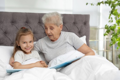 Cute girl and her grandmother reading book on bed at home