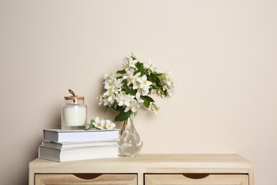 Photo of Bouquet of beautiful jasmine flowers in glass vase, books and candle on wooden commode near beige wall indoors