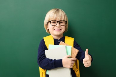 Photo of Happy little school child with notebooks showing thumbs up near chalkboard