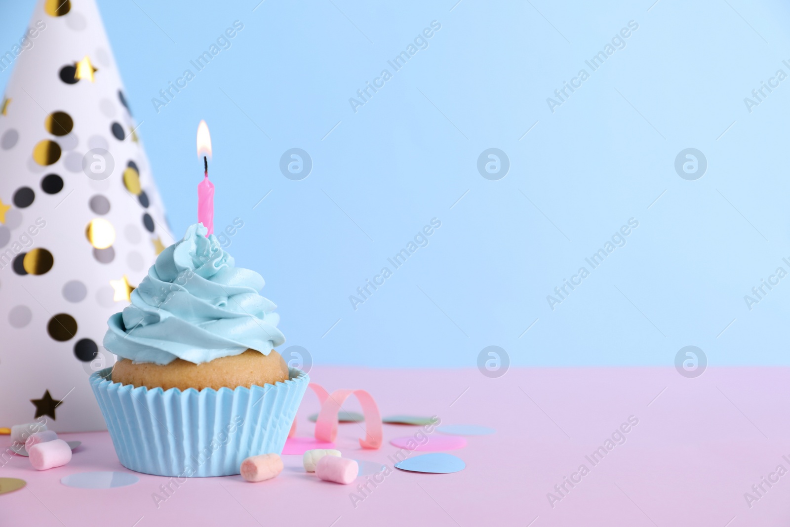 Photo of Delicious birthday cupcake with burning candle, marshmallows and party decor on pink table against light blue background, space for text