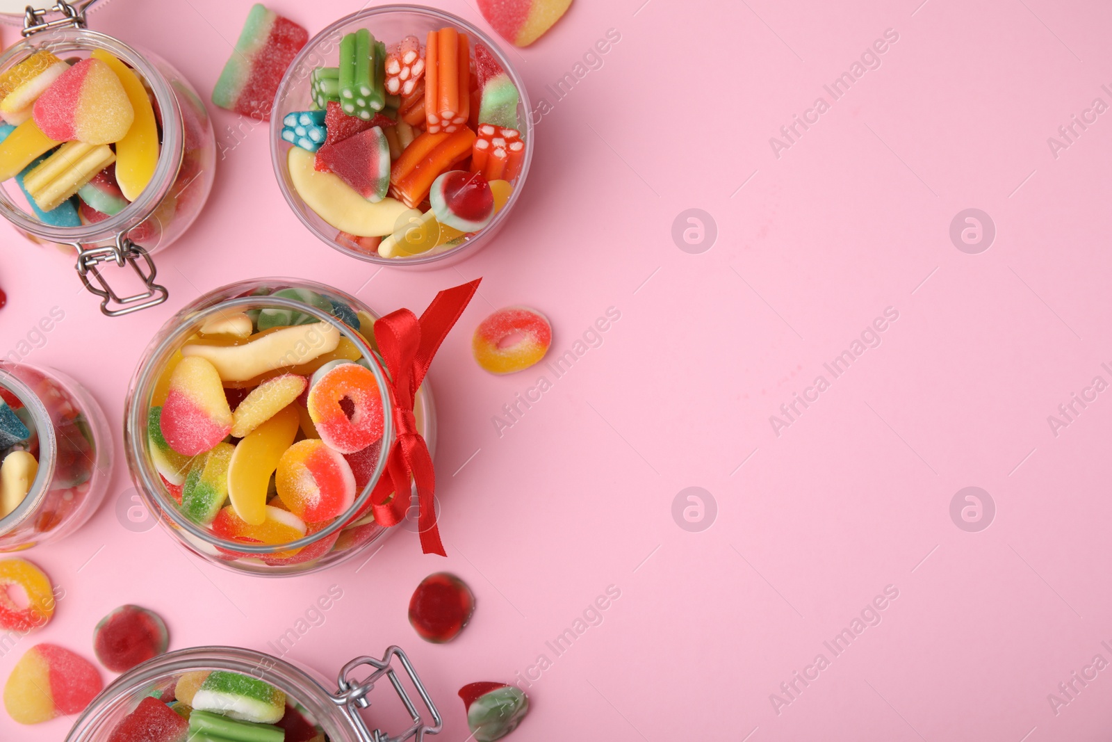 Photo of Glass jars with tasty colorful jelly candies on pink background, flat lay. Space for text