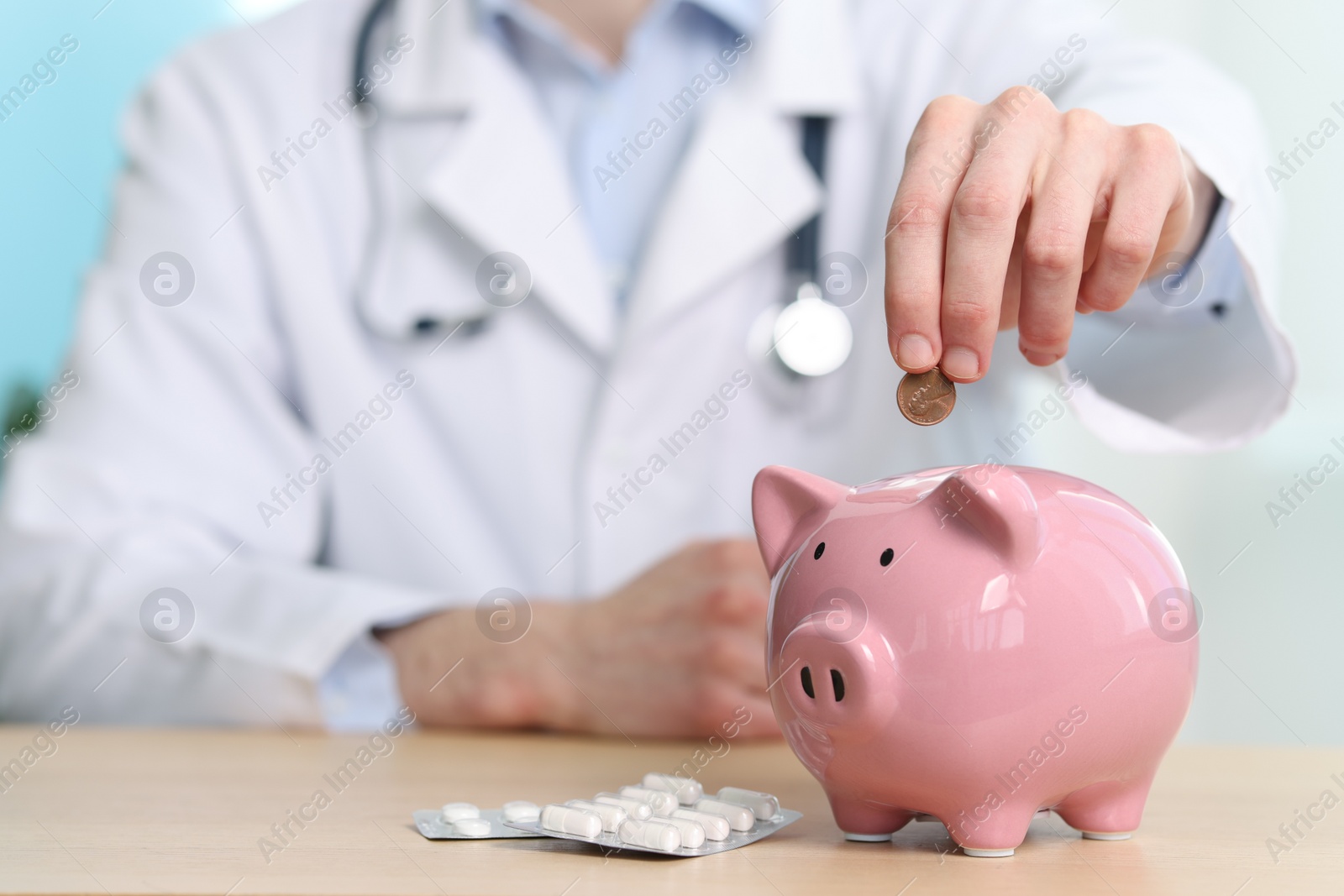 Photo of Doctor putting coin into piggy bank at wooden table, closeup