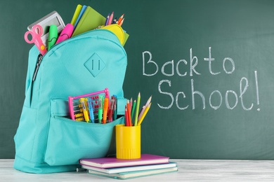 Bright backpack with school stationery on wooden table near green chalkboard. Back to School