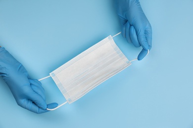 Doctor in medical gloves holding protective mask on light blue background, closeup. Space for text