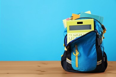 Backpack with different school stationery on wooden table against light blue background, space for text
