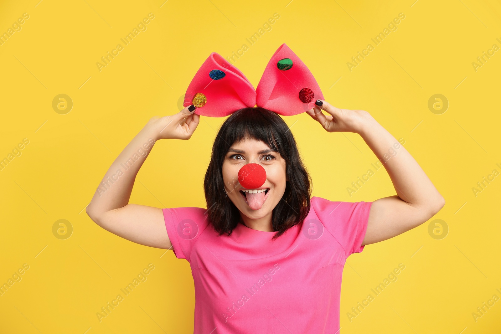 Photo of Funny woman with large bow and clown nose on yellow background. April fool's day