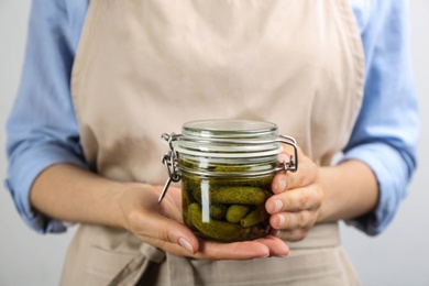 Woman holding glass jar of pickled cucumbers on light background, closeup