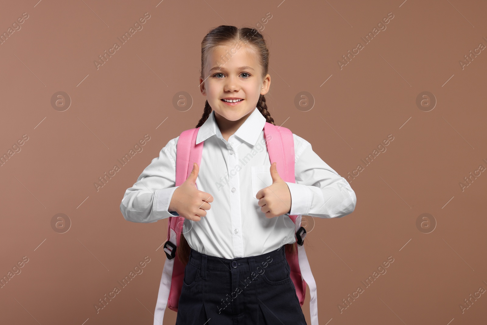 Photo of Happy schoolgirl with backpack showing thumbs up gesture on brown background