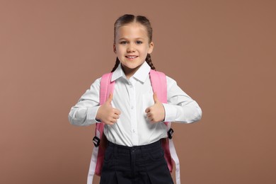 Photo of Happy schoolgirl with backpack showing thumbs up gesture on brown background