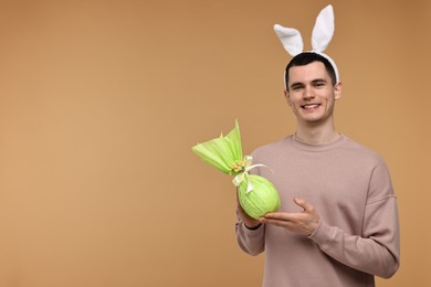 Photo of Easter celebration. Handsome young man with bunny ears holding wrapped gift on beige background. Space for text