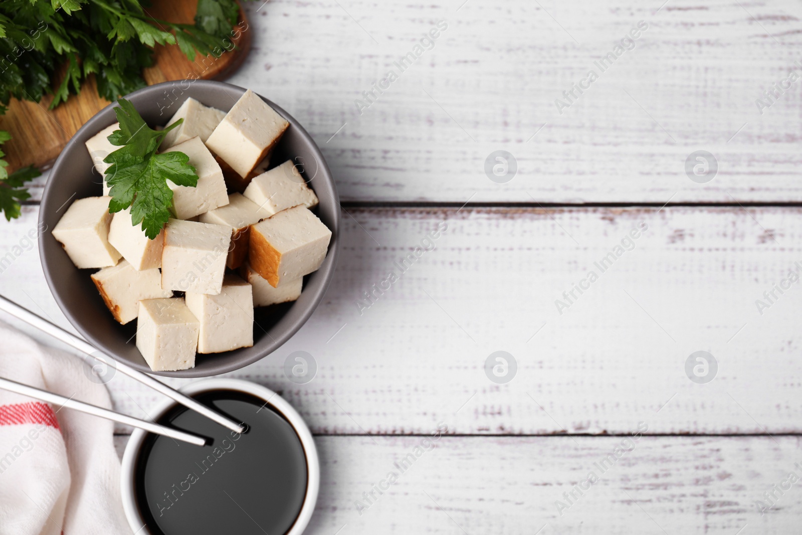 Photo of Bowl of smoked tofu cubes, soy sauce and parsley on white wooden table, flat lay. Space for text