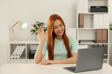 Young woman waving hello during video chat via laptop at table in office