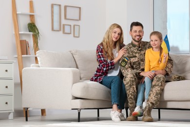 Soldier in military uniform reunited with his family and Ukrainian flag on sofa at home, space for text