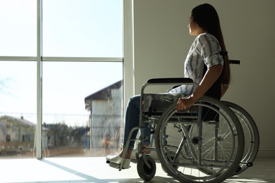 Photo of Young woman in wheelchair near window indoors. Space for text