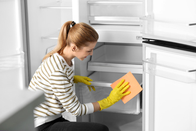 Photo of Woman in rubber gloves cleaning empty refrigerator with rag at home