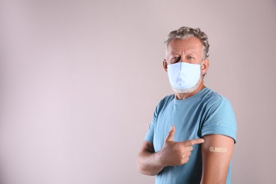Senior man in protective mask pointing at arm with bandage after vaccination on beige background. Space for text