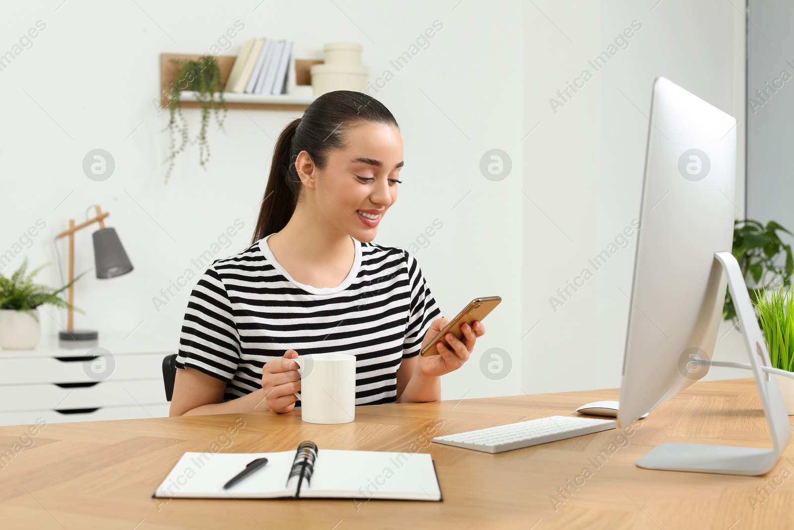 Photo of Home workplace. Happy woman with cup of hot drink looking at smartphone at wooden desk in room