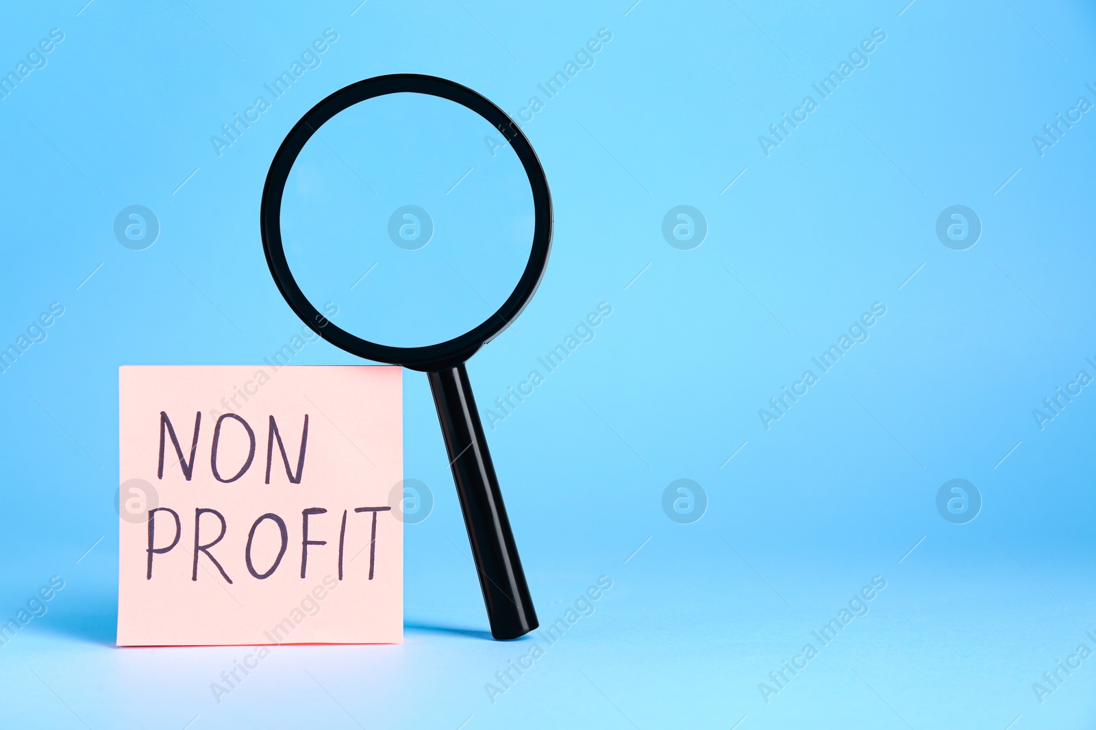 Photo of Paper note with phrase Non Profit and magnifying glass on light blue background. Space for text