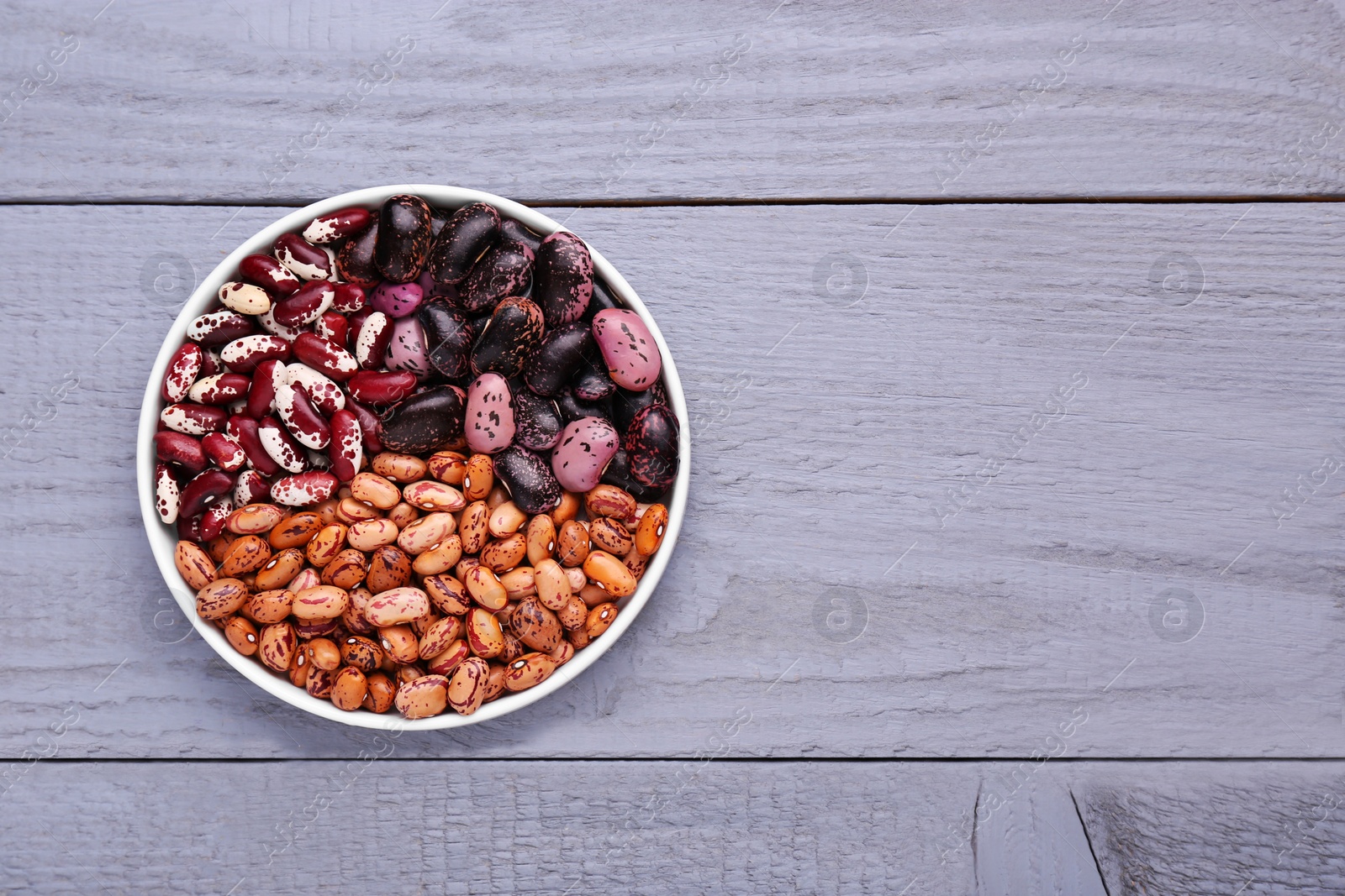 Photo of Different kinds of dry kidney beans in bowl on light grey wooden table, top view. Space for text