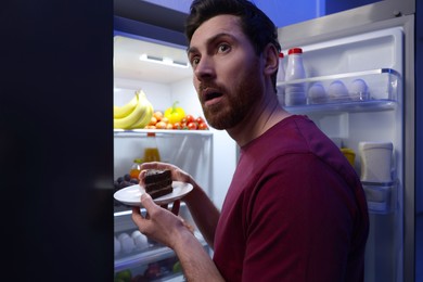 Man taking plate with cake from refrigerator in kitchen at night. Bad habit
