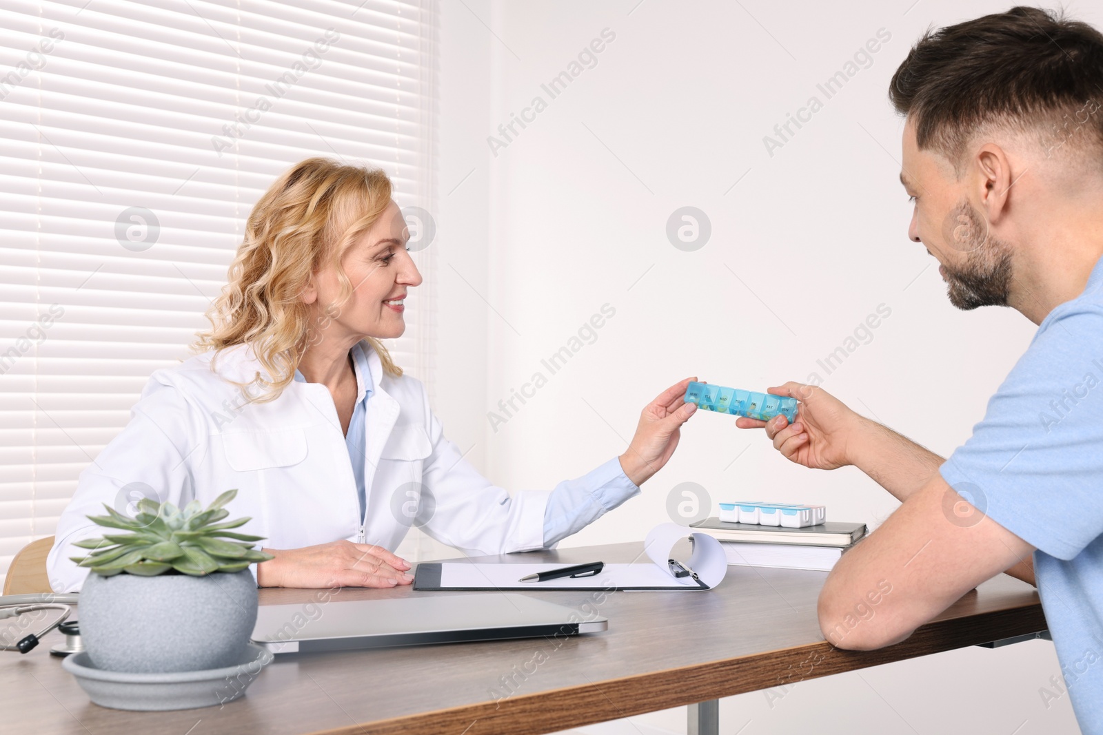 Photo of Doctor holding box of pills and consulting patient at table in clinic