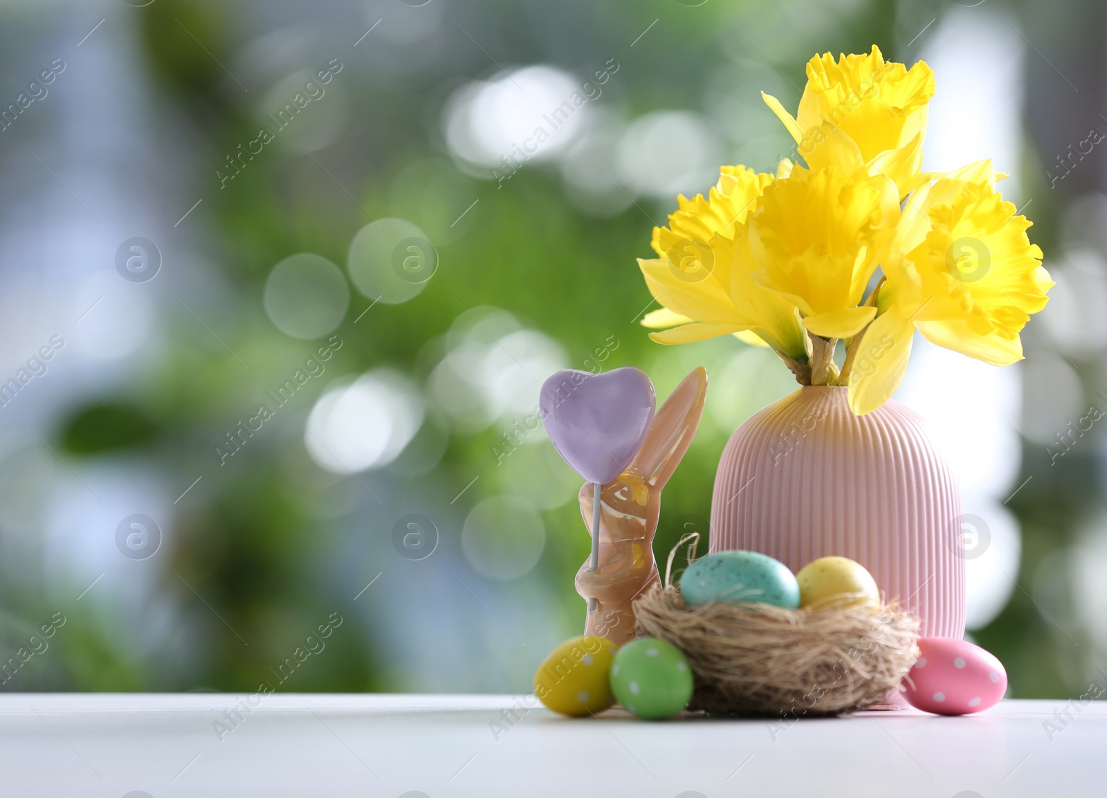 Photo of Painted Easter eggs, ceramic bunny and flowers on white table against blurred background. Space for text