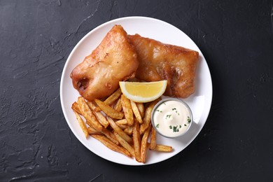 Photo of Tasty fish, chips, lemon and sauce on black table, top view