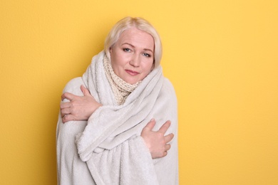 Mature woman wrapped in blanket suffering from cold on color background