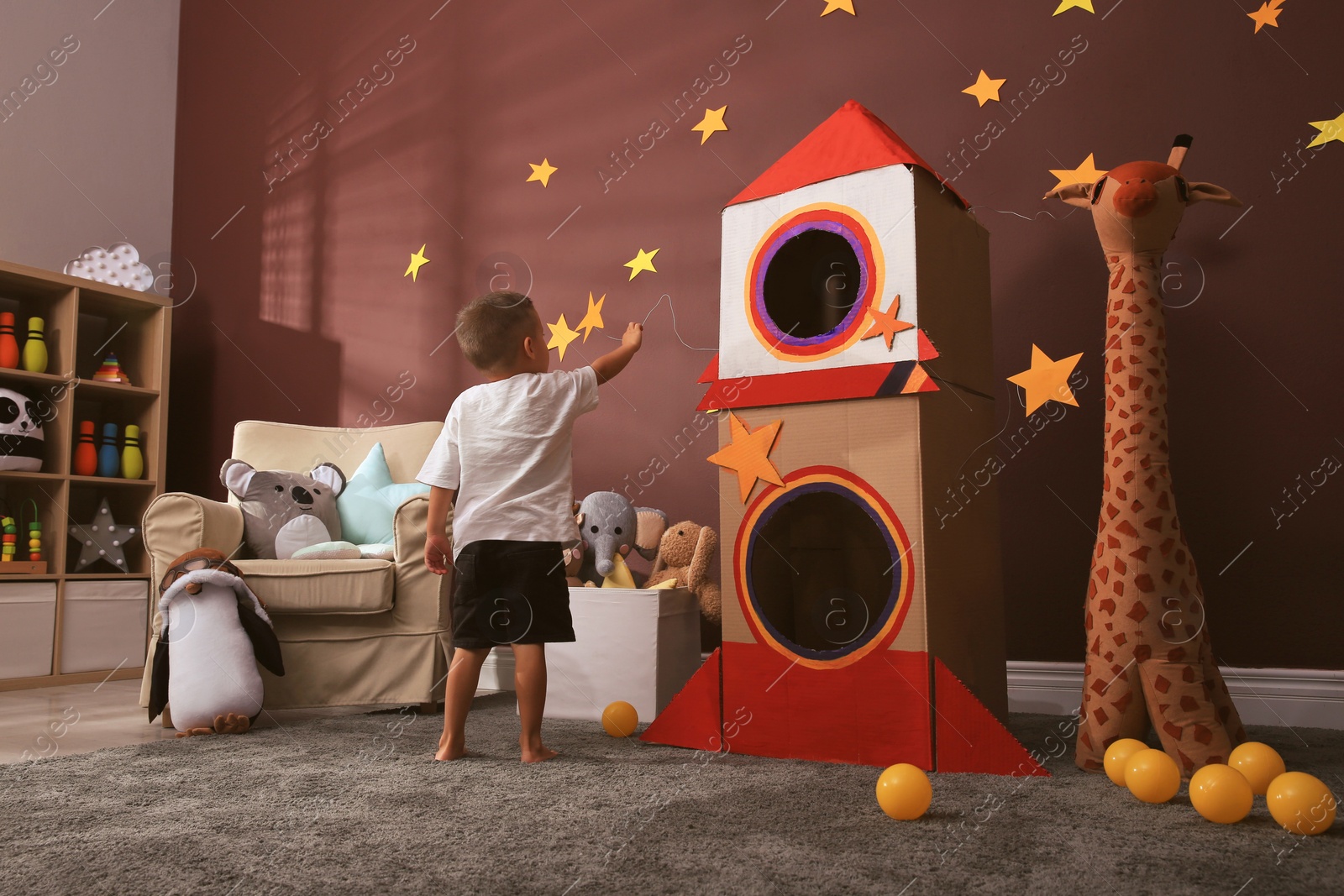 Photo of Cute little boy playing on floor near cardboard rocket  at home. Child's room interior