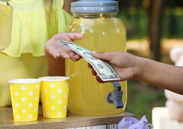 Photo of Little girl selling natural lemonade to African-American boy in park, closeup. Summer refreshing drink