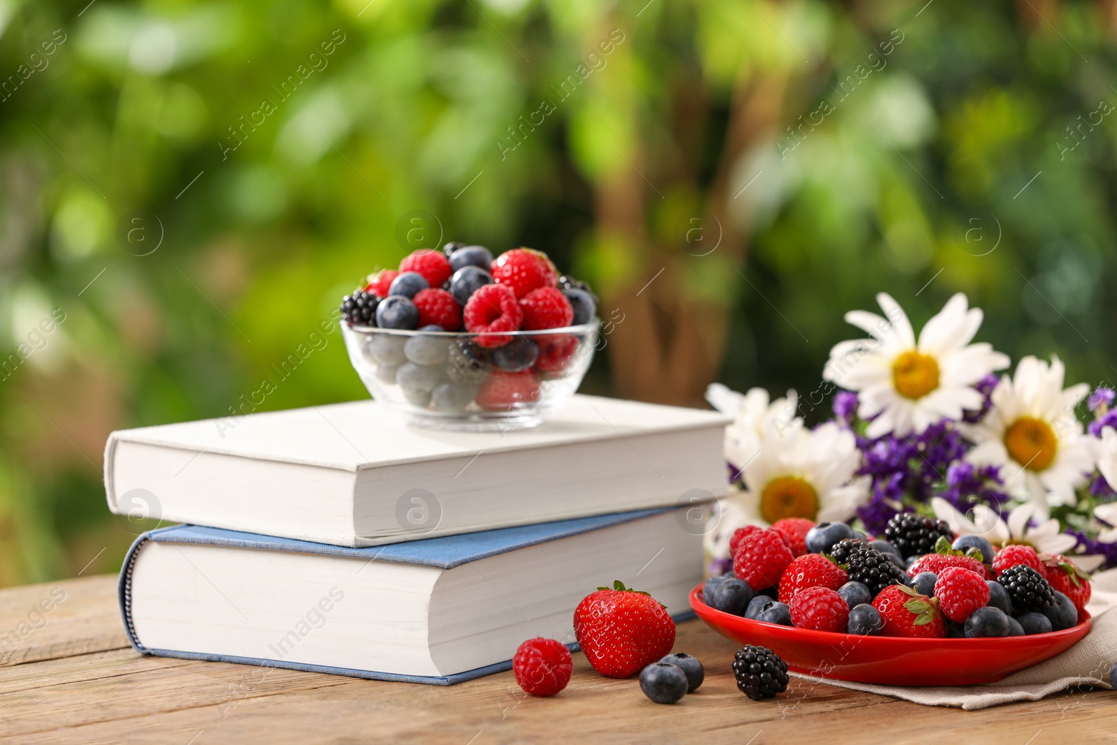 Photo of Different fresh ripe berries, beautiful flowers and books on wooden table outdoors