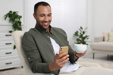 Photo of Happy man with cup of drink sending message via smartphone at home