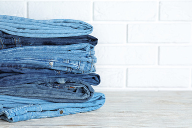 Photo of Stack of different jeans on white wooden table against brick wall. Space for text