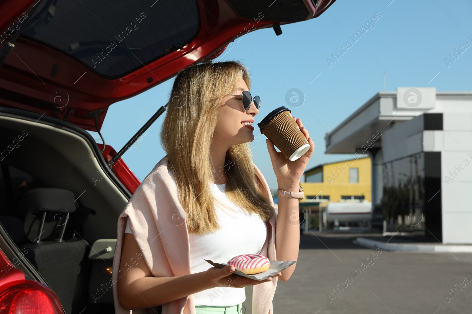 Photo of Beautiful young woman with doughnut drinking coffee near car at gas station