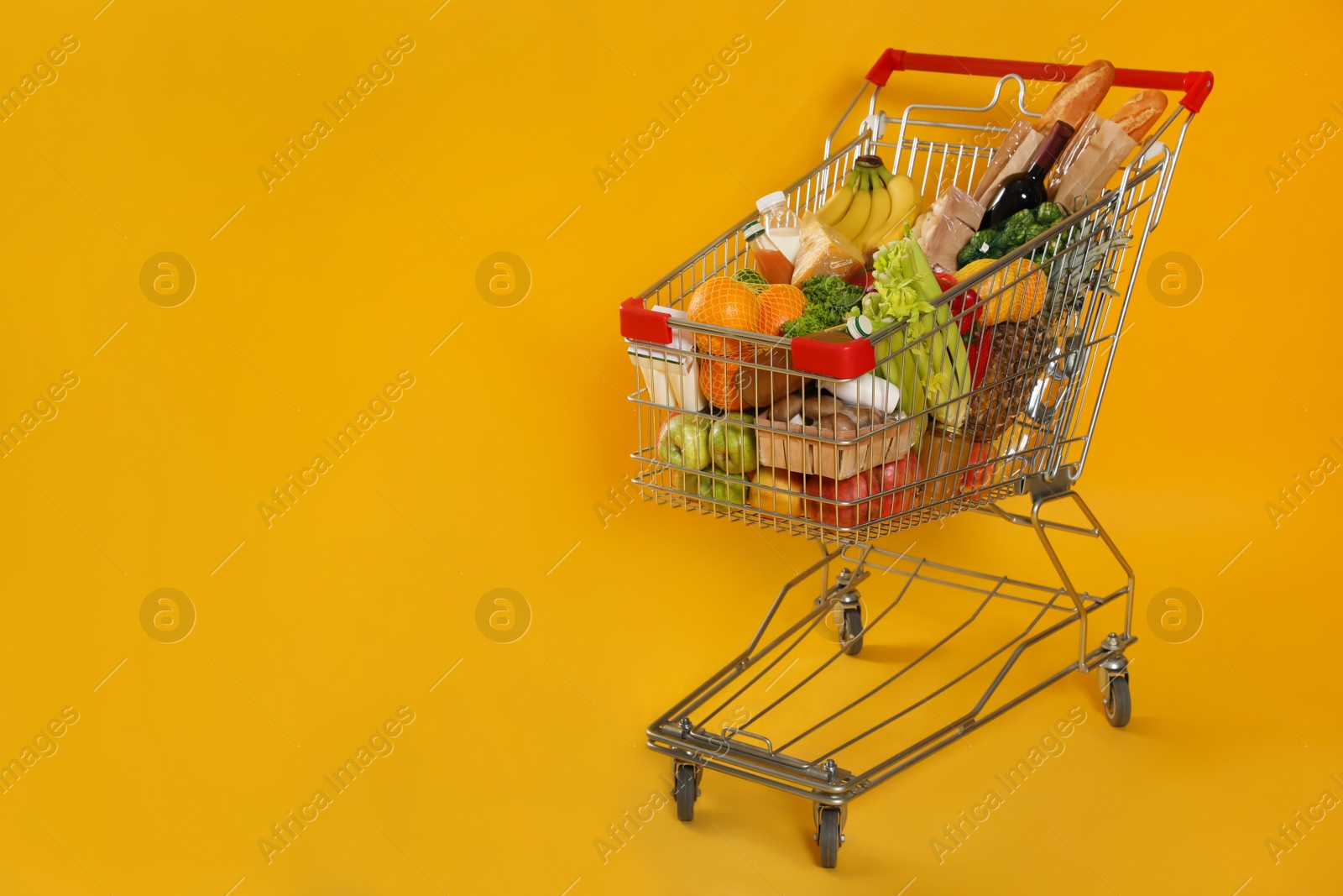 Photo of Shopping cart full of groceries on yellow background. Space for text