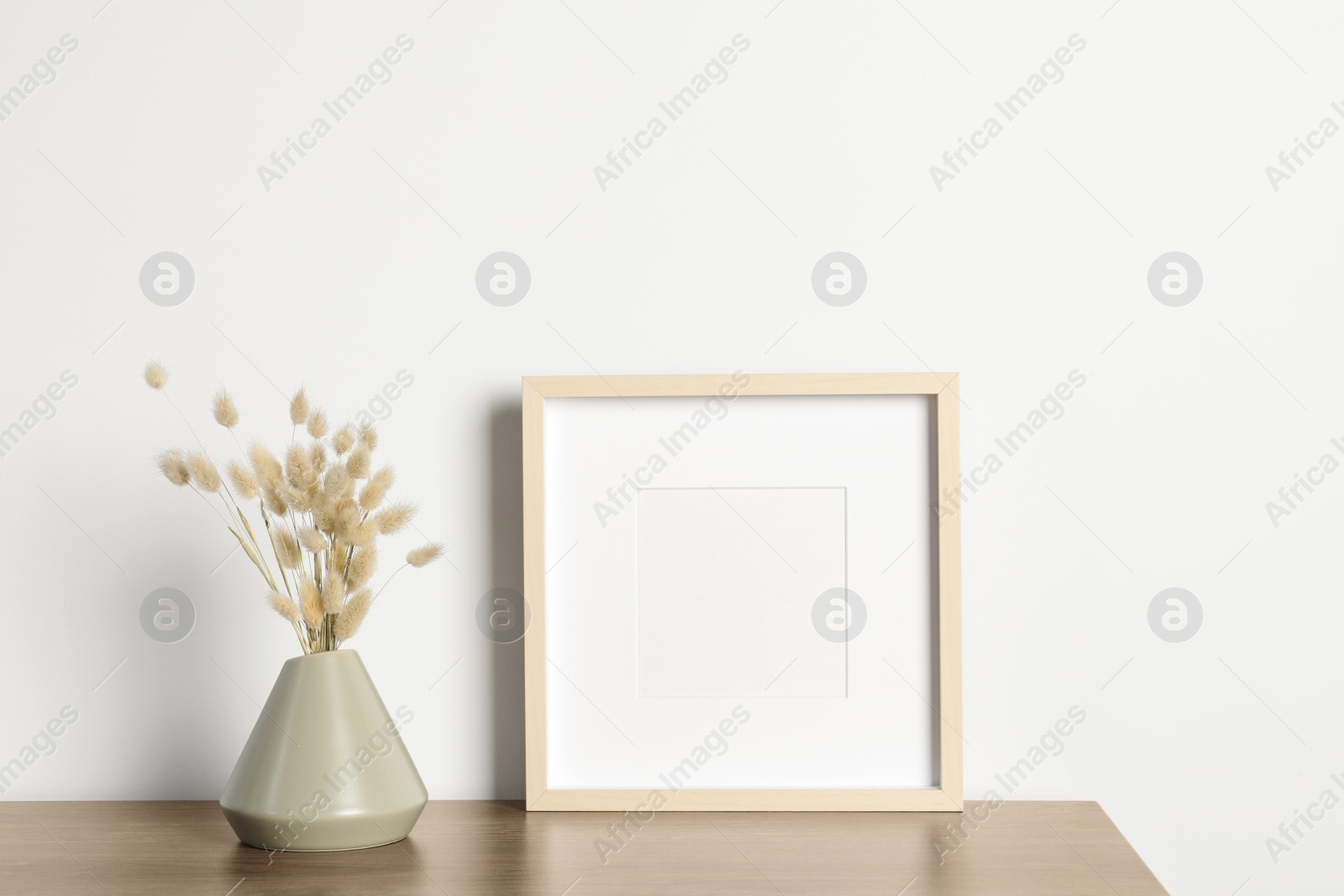 Photo of Empty photo frame and vase with dry decorative spikes on wooden table. Mockup for design