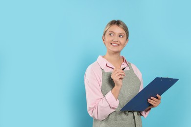 Photo of Beautiful young woman in clean apron with clipboard on light blue background. Space for text