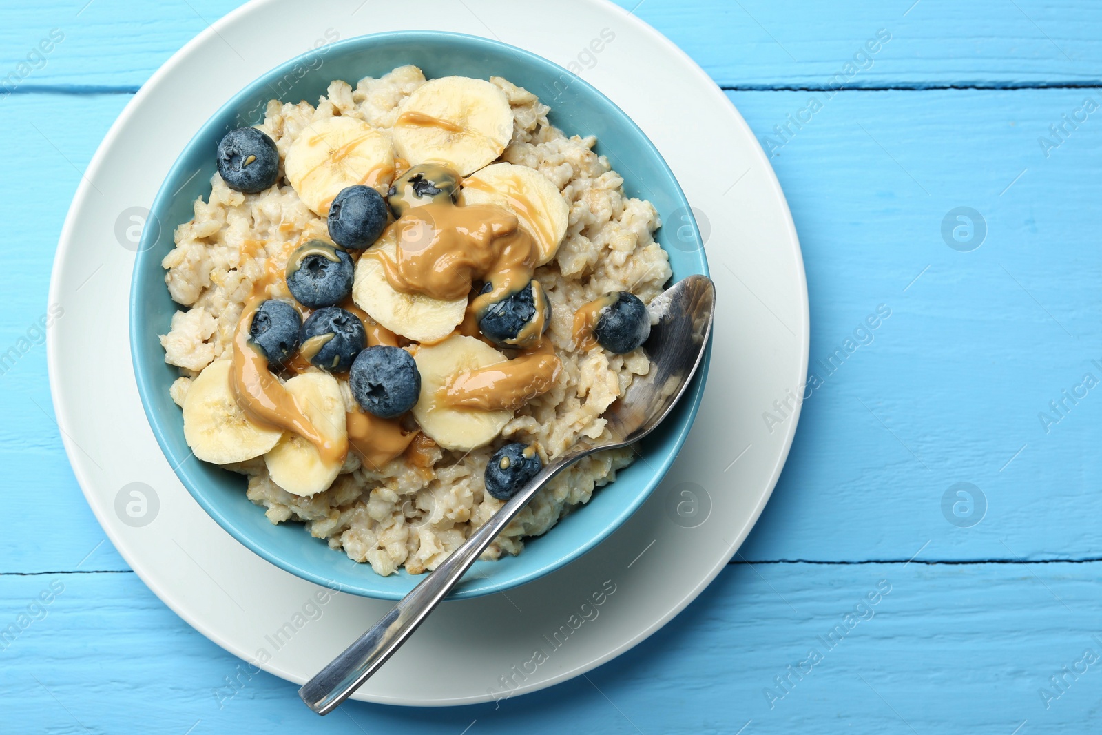 Photo of Tasty oatmeal with banana, blueberries and peanut butter served in bowl on light blue wooden table, top view. Space for text