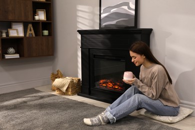 Photo of Beautiful young woman with cup of hot drink sitting on floor near fireplace at home. Space for text