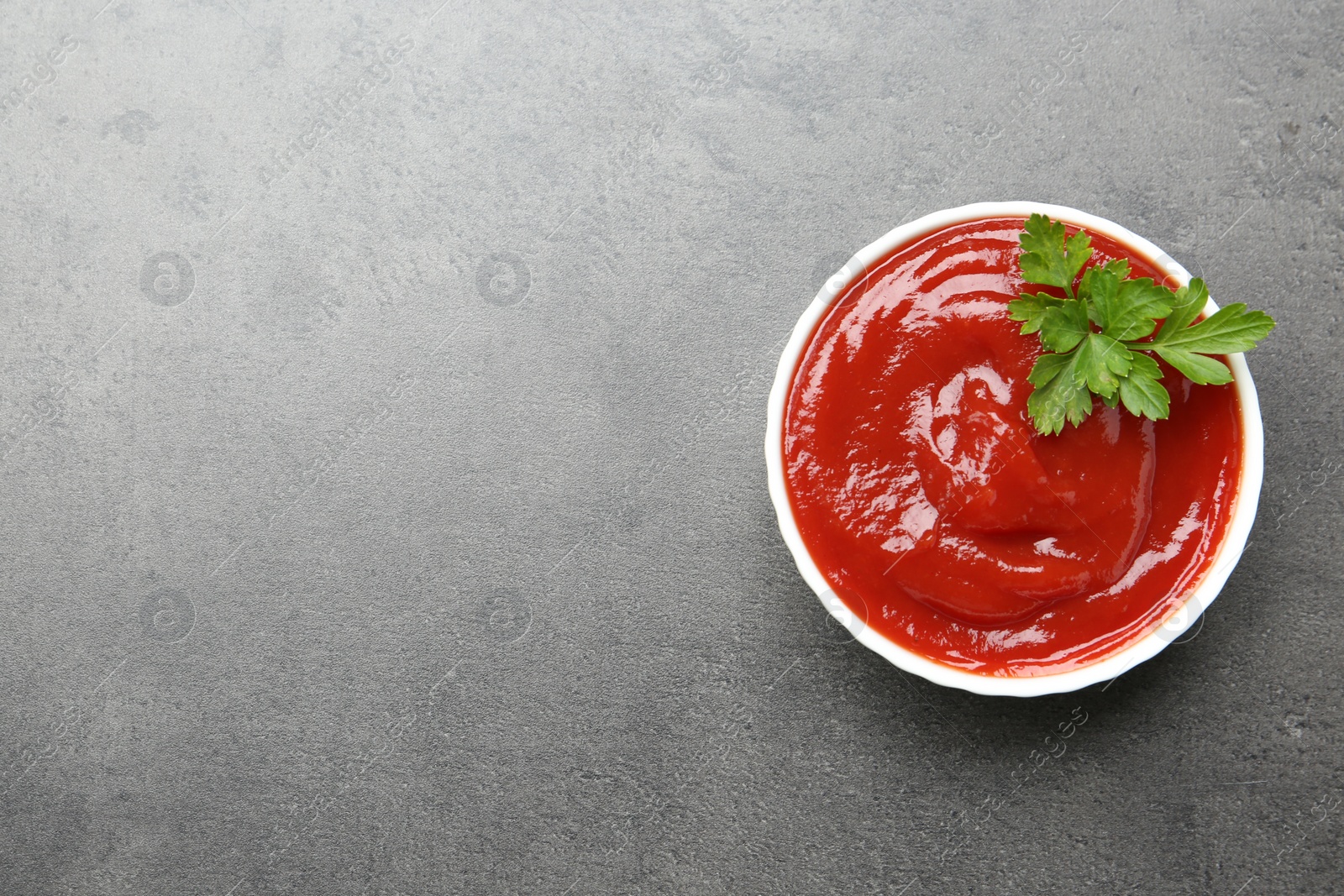 Photo of Delicious tomato ketchup and parsley in bowl on grey textured table, top view. Space for text