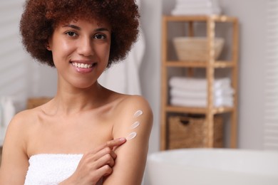 Beautiful young woman applying body cream onto arm in bathroom, space for text