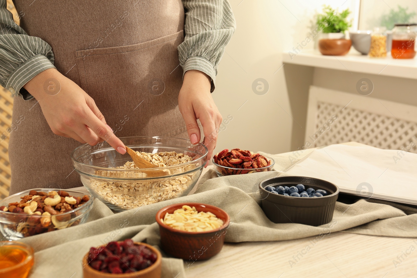 Photo of Making granola. Woman putting oat flakes into bowl at table in kitchen, closeup