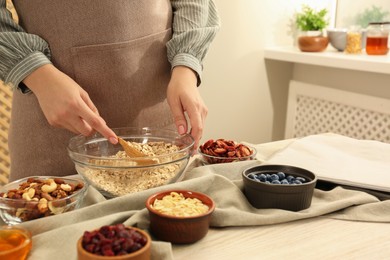 Photo of Making granola. Woman putting oat flakes into bowl at table in kitchen, closeup