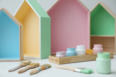 Photo of House shaped shelves, jars of paints and brushes on white table. Interior elements