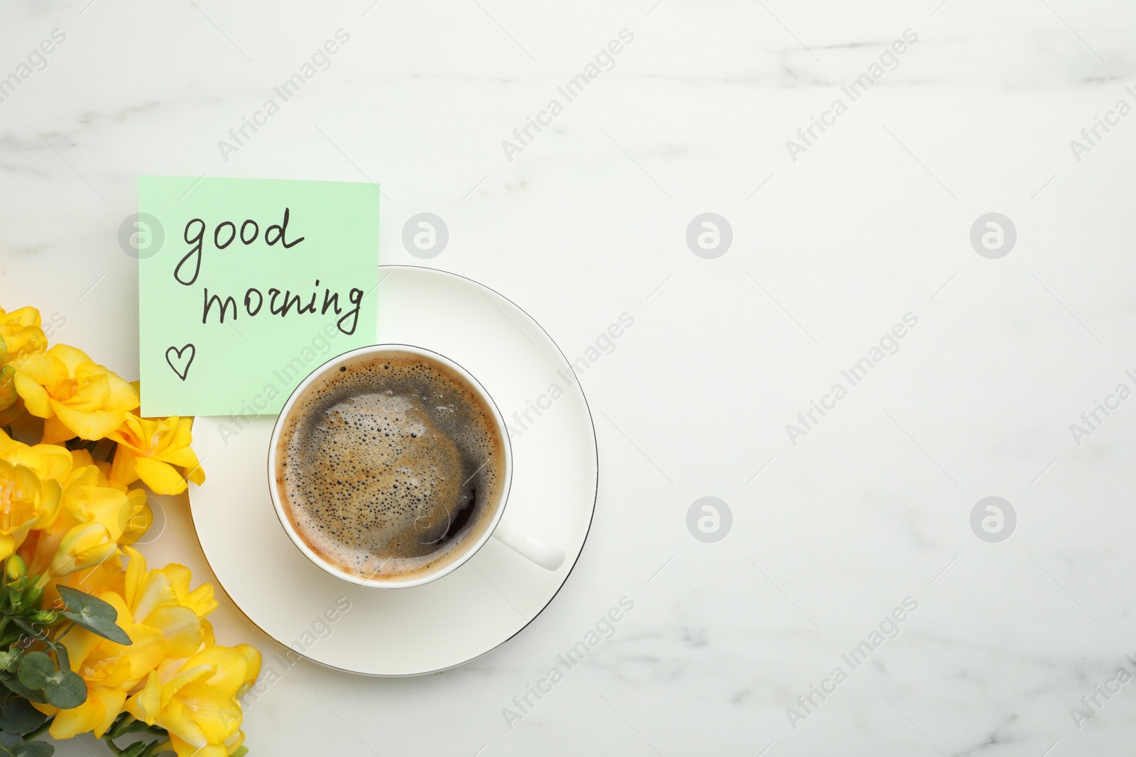 Photo of Cup of aromatic coffee, beautiful yellow freesias and Good Morning note on white marble table, flat lay. Space for text