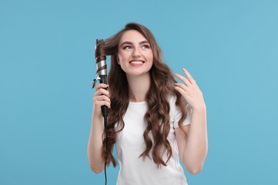 Photo of Beautiful young woman using curling hair iron on light blue background