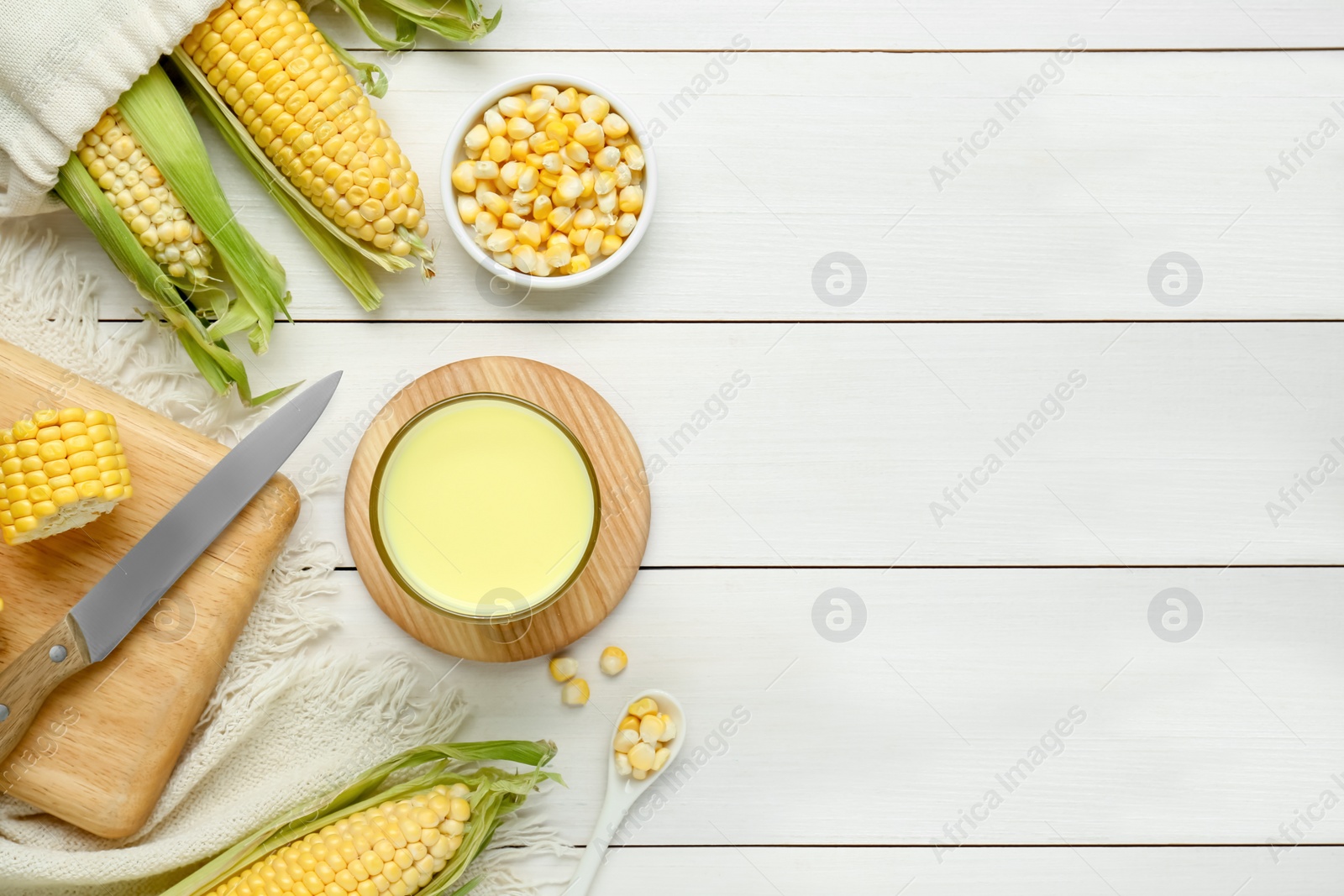 Photo of Tasty fresh corn milk in glass and cobs on white wooden table, flat lay. Space for text