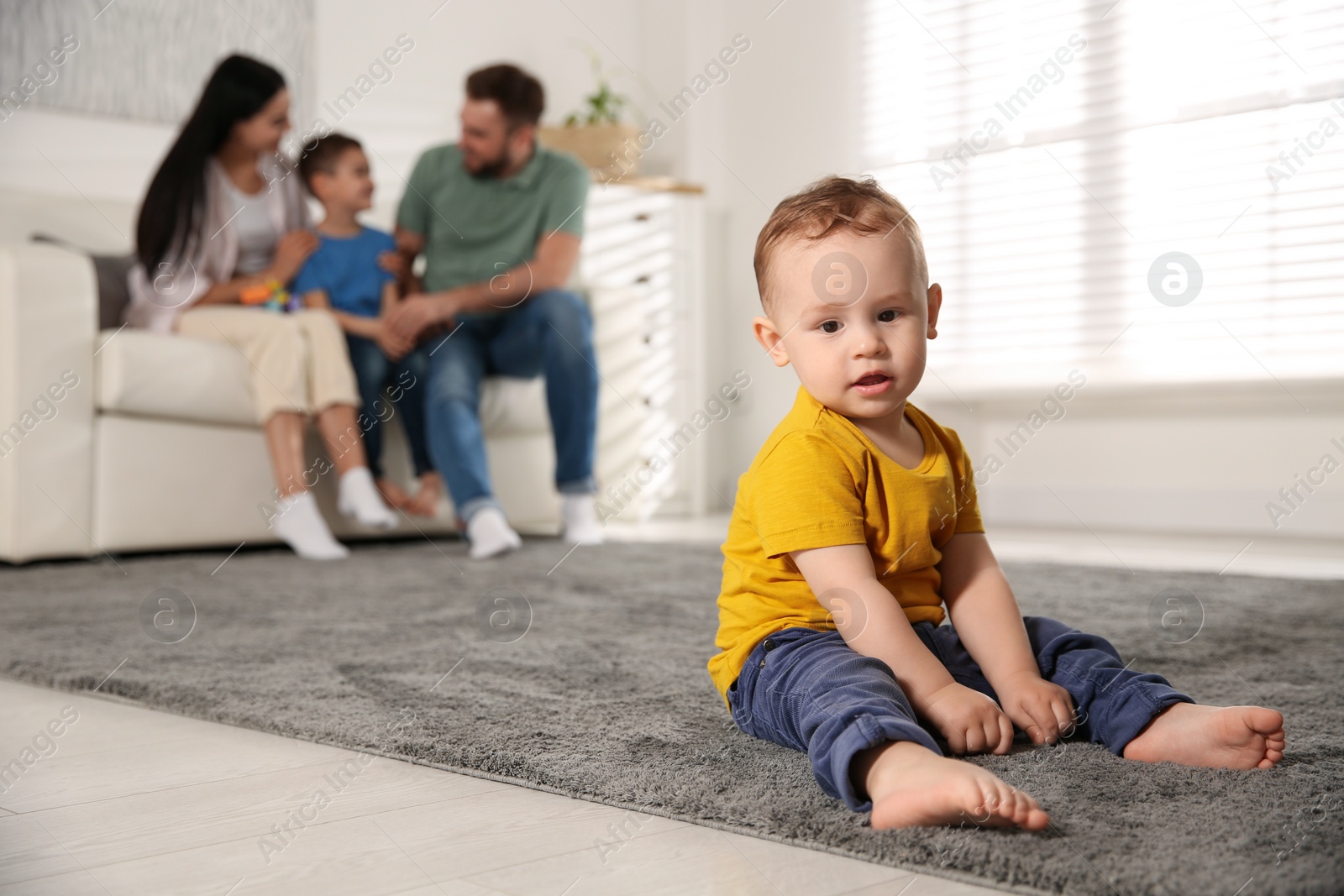 Photo of Unhappy baby sitting alone on floor while parents spending time with his elder brother at home. Jealousy in family